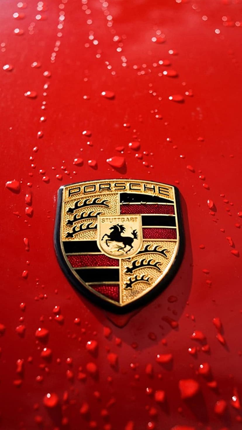 Porsche crest emblem centered on a red car hood covered in small water droplets during a rain shower.