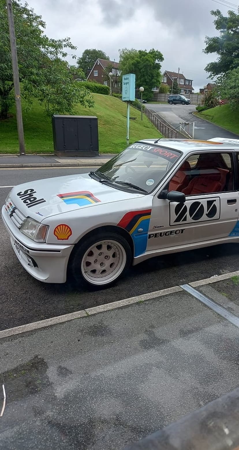 White Peugeot 205 GTi rally car featuring Shell decals sits on wet asphalt under a gray overcast sky.
