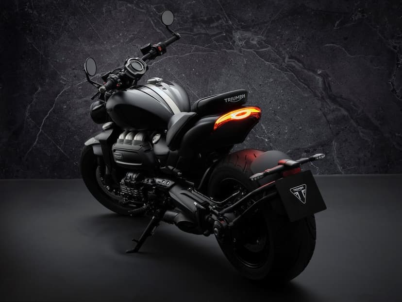 Rear view of a matte black Triumph Rocket 3 motorcycle with illuminated taillights against a dark slate wall.
