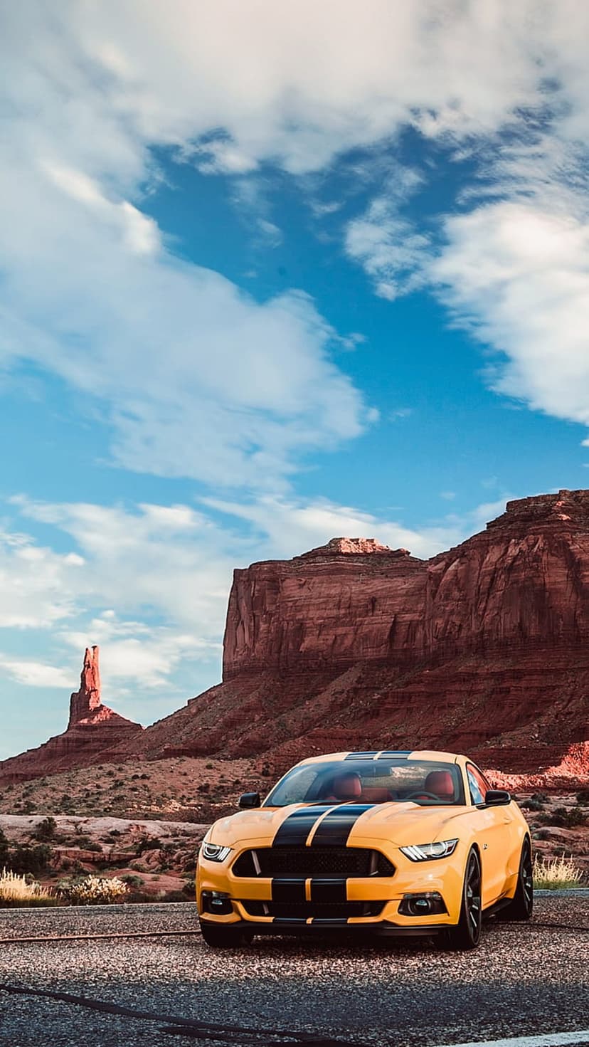 Yellow Ford Mustang with black stripes on a desert road facing red rock buttes under a blue sky in Monument Valley.