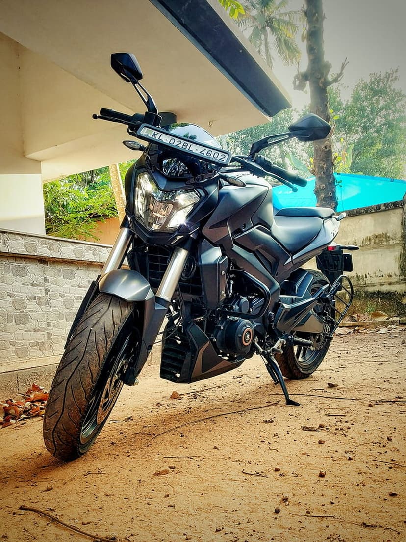 Black sport bike with sharp fairings sits on a dirt ground under a shaded structure near green foliage.