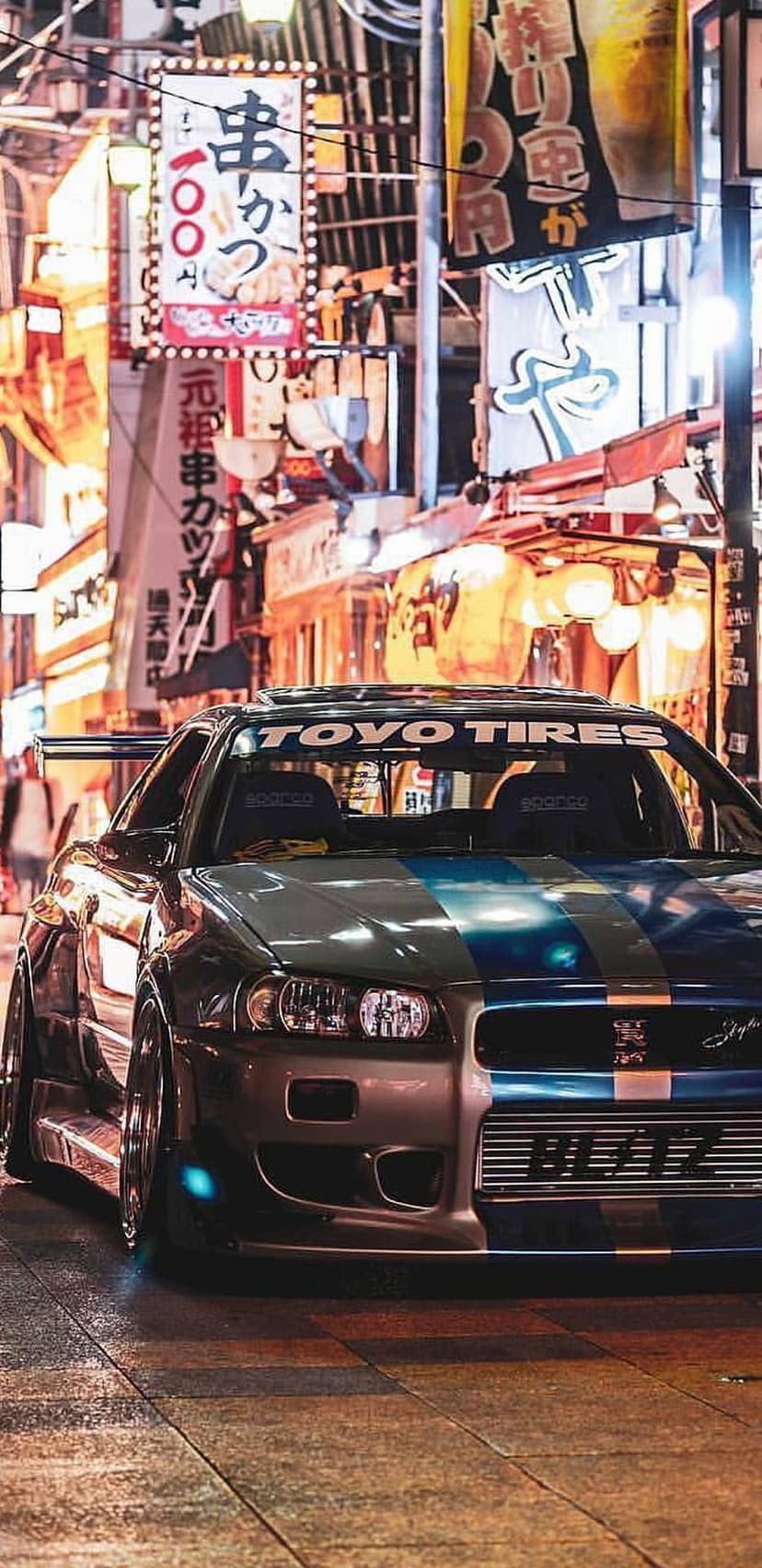 Nissan Skyline R34 parked on a rainy Tokyo street with glowing neon signs and traditional Japanese lanterns.