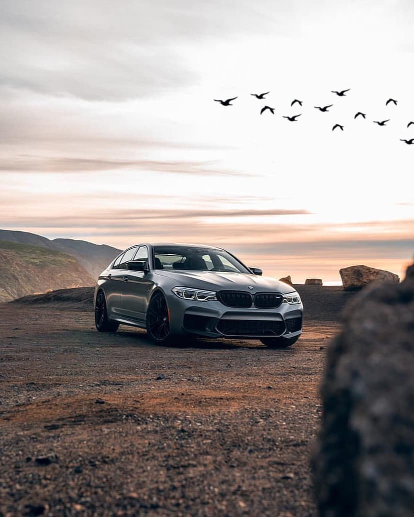 BMW M5 sedan parked on a rocky cliff edge above the sea with birds flying through a purple and orange sunset sky.