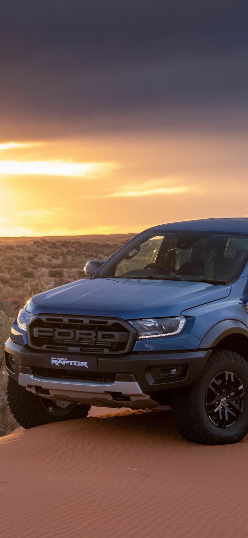 Blue Ford Ranger Raptor truck on a red sand dune under a golden sunset sky in a vast desert landscape.