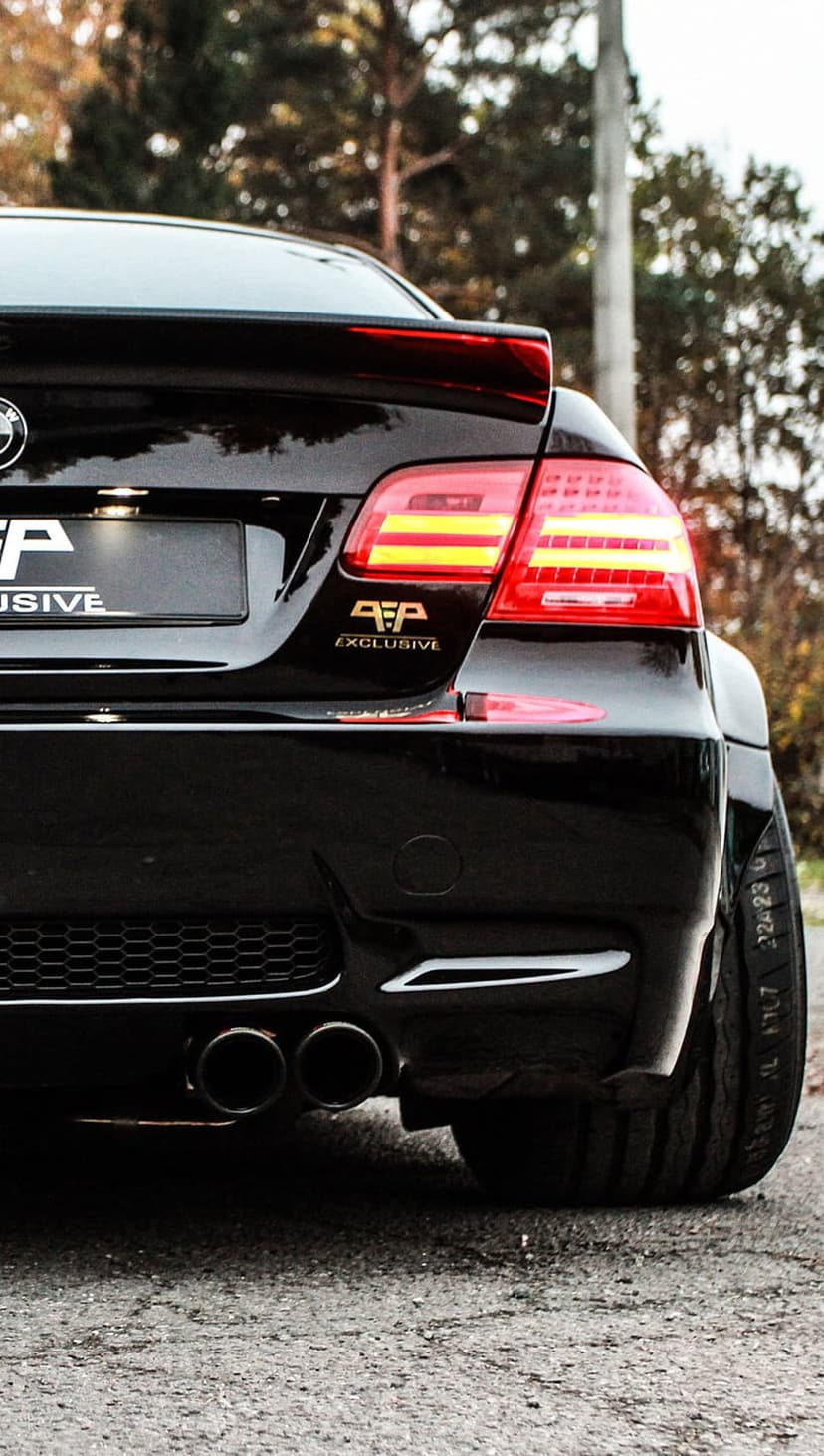 BMW M3 rear view featuring black paint, dual exhausts, a spoiler, and glowing red LED taillights outdoors.
