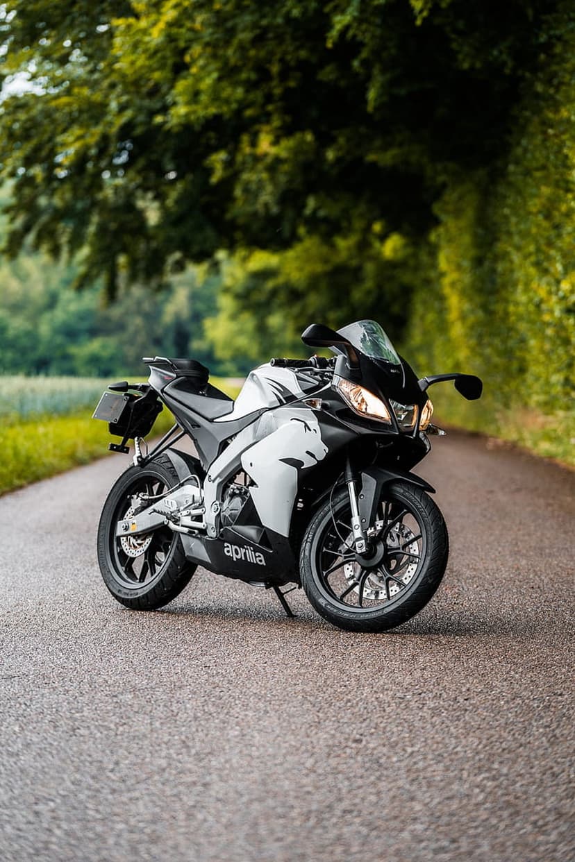 Aprilia sportbike with black and white graphics parked on a wet asphalt road flanked by lush green trees.