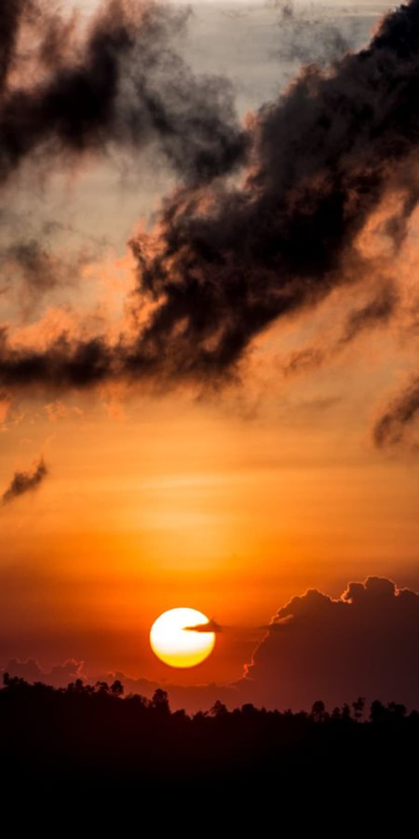 Radiant orange sun setting behind dark hill silhouettes with dramatic charcoal clouds in a vertical sky view.