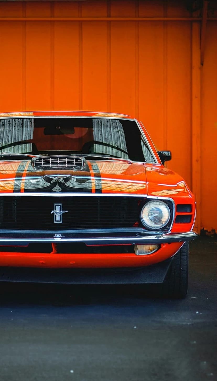 Orange Ford Mustang with black racing stripes parked against a matching textured wall in a low-angle vertical shot.