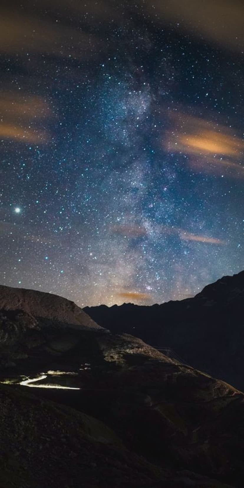 Milky Way galaxy arches over silhouetted mountains and a glowing winding road under a dark, starry night sky.