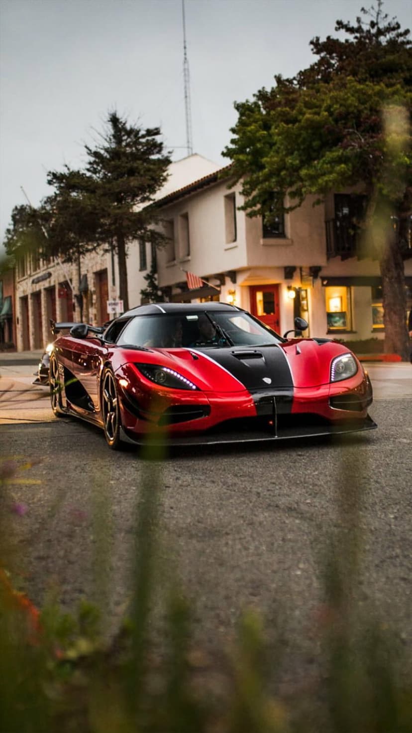 Koenigsegg hypercar with a red and black exterior parked on a city street beside trees and modern buildings.