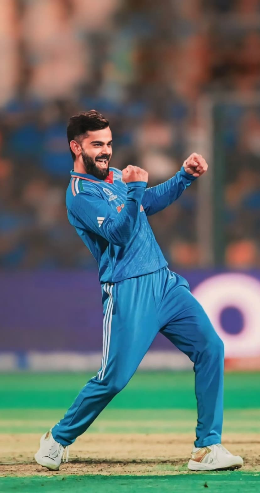 Virat Kohli raises his fists in triumph while wearing a blue cricket jersey on a blurred grass field background.
