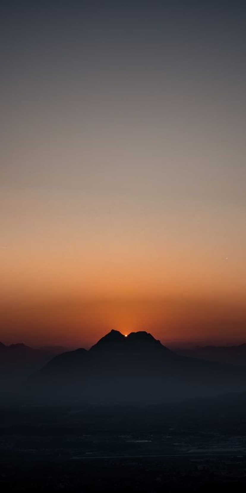 Mountain peaks silhouette against a gradient orange and grey sky as the sun dips behind a sharp rocky ridge.