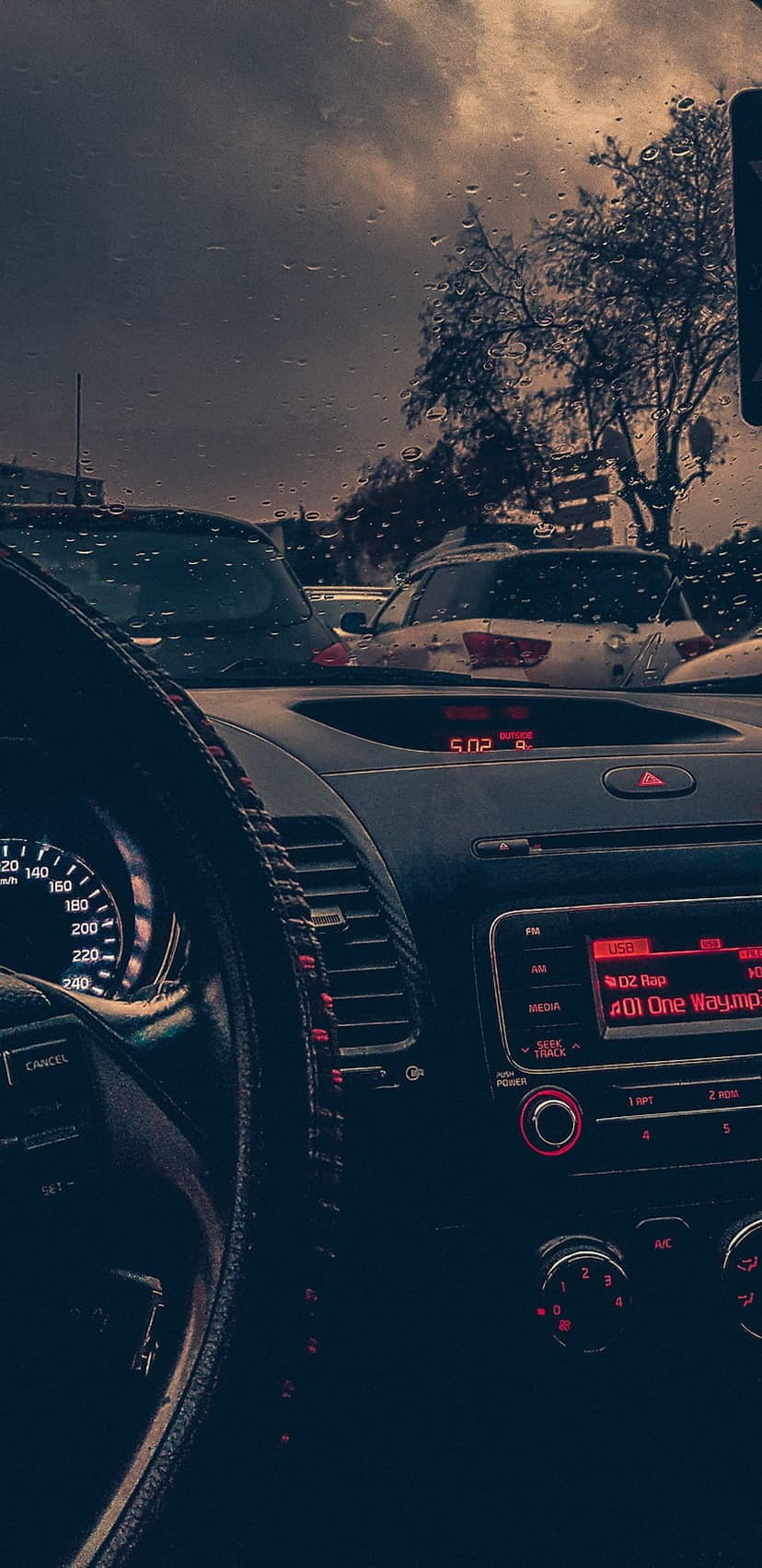 Raindrops on a car windshield overlook a dark steering wheel and dashboard with glowing red interior lights.