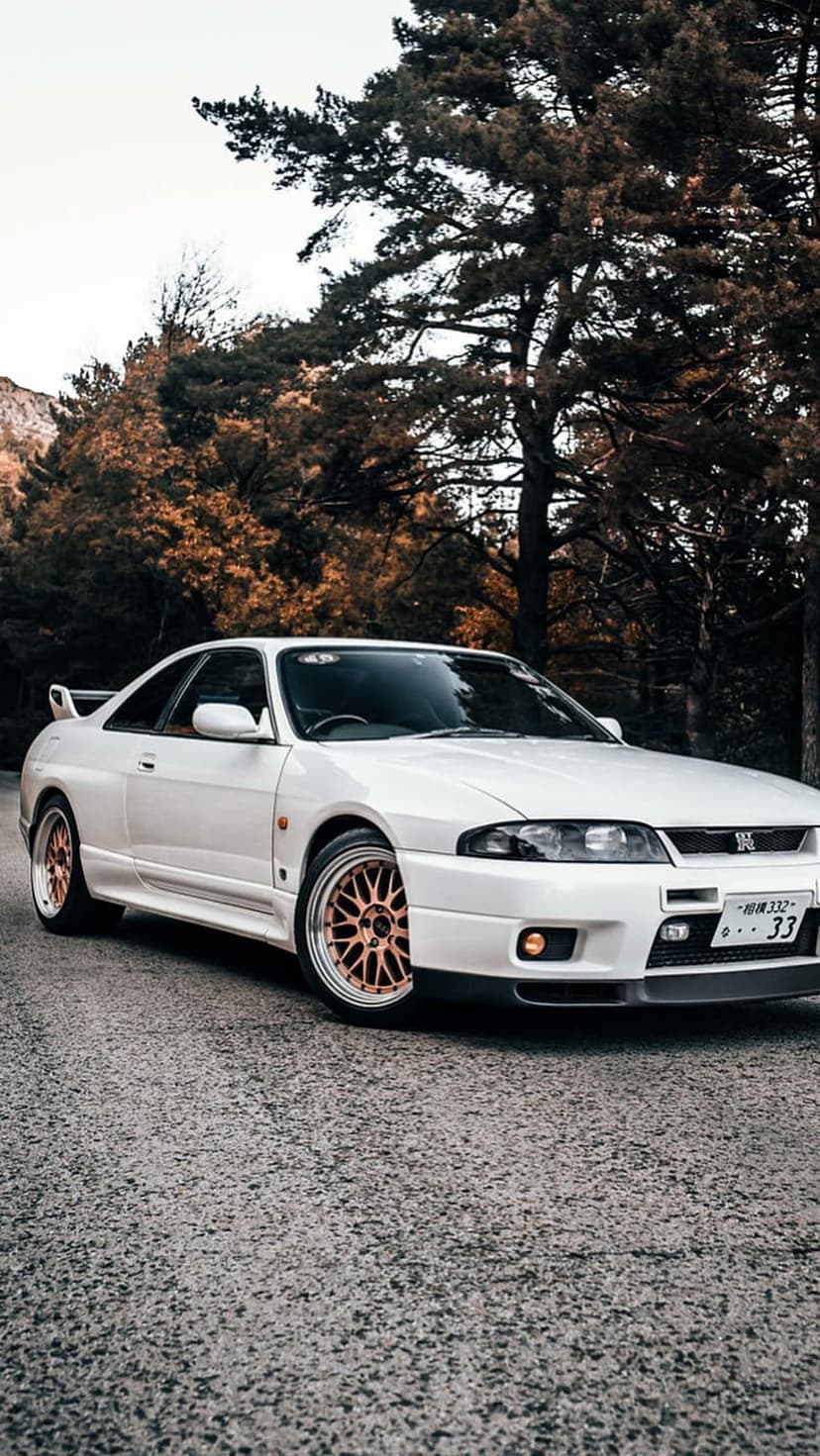 White Nissan Skyline R33 GT-R with gold rims on a paved road surrounded by orange autumn trees under a grey sky.