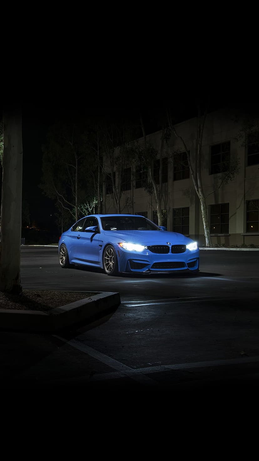 Blue BMW M4 coupe with glowing headlights parked on a dark asphalt road in front of a building at night.