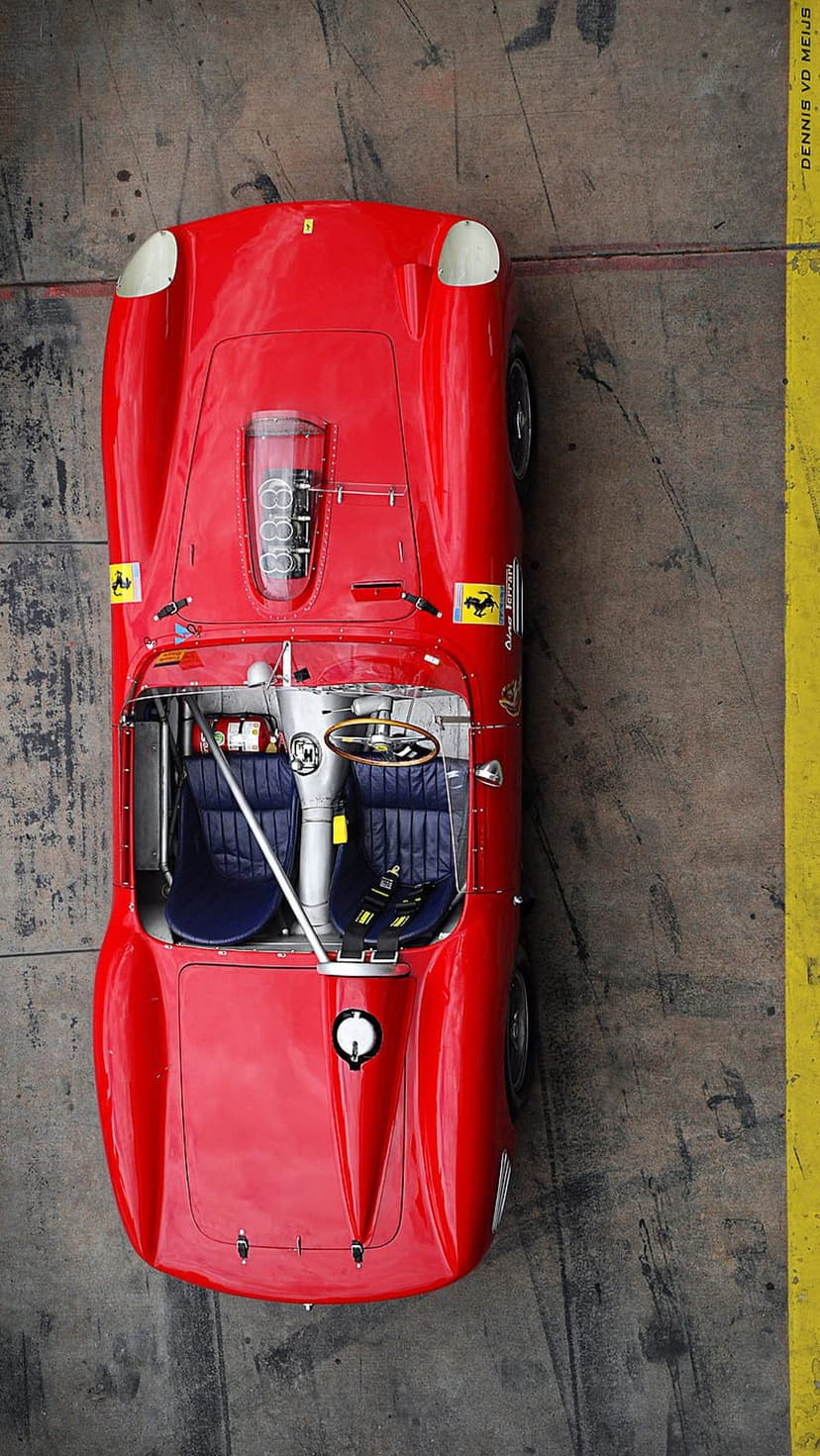 Red Ferrari race car cockpit with navy leather seats and wooden steering wheel on a concrete track surface.