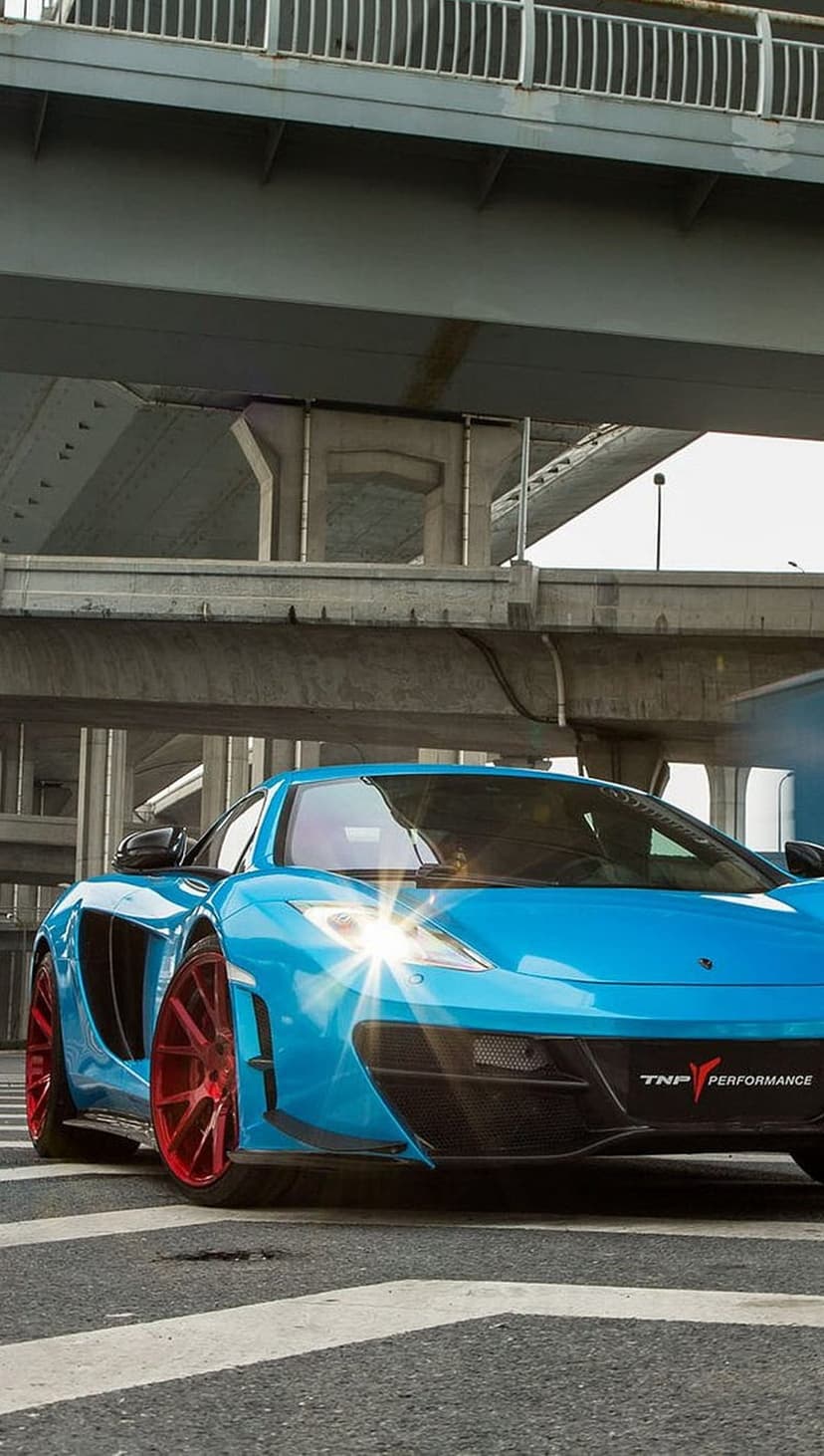 Blue McLaren supercar with red wheels parked on a crosswalk under a concrete bridge with sunlight on headlights.