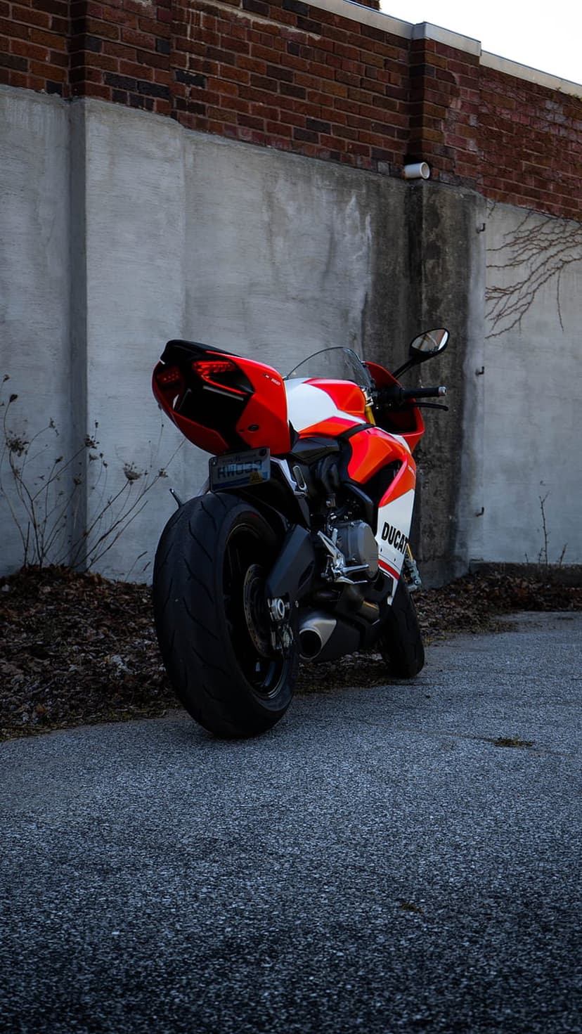 Ducati motorcycle rear view parked on asphalt against a textured concrete wall with a brick upper facade.