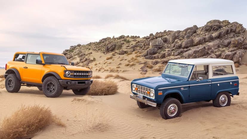 Classic blue and modern yellow Ford Broncos on desert sand dunes with rugged rock hills in the background.
