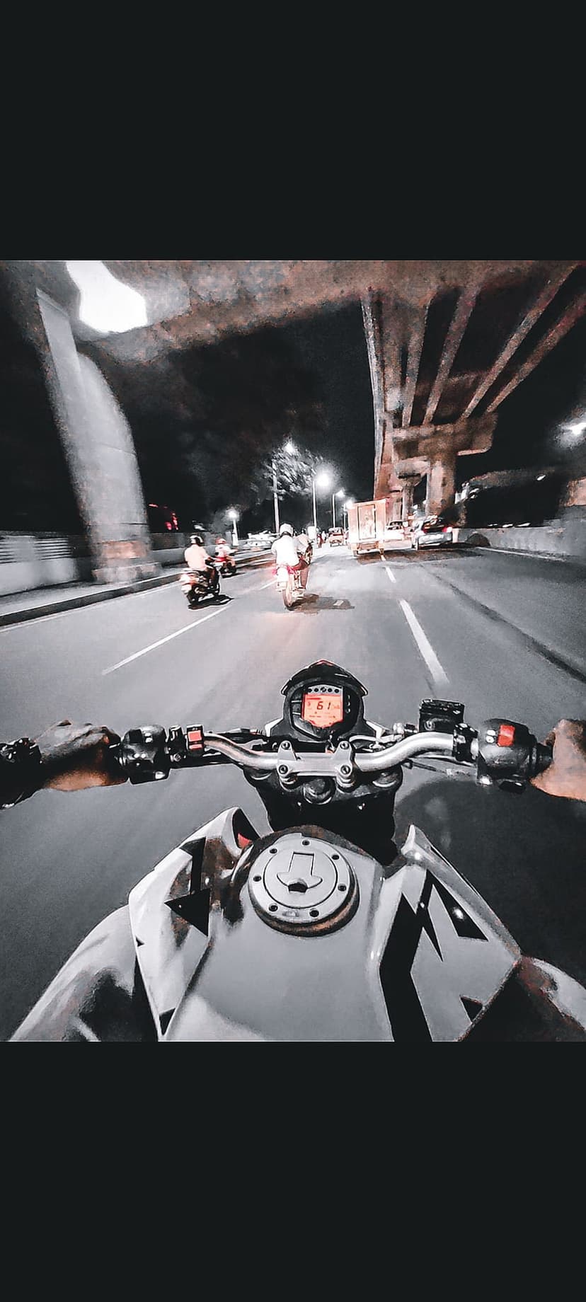 Motorcycle handlebars and digital speedometer seen from the rider's POV on a city highway at night under an overpass.