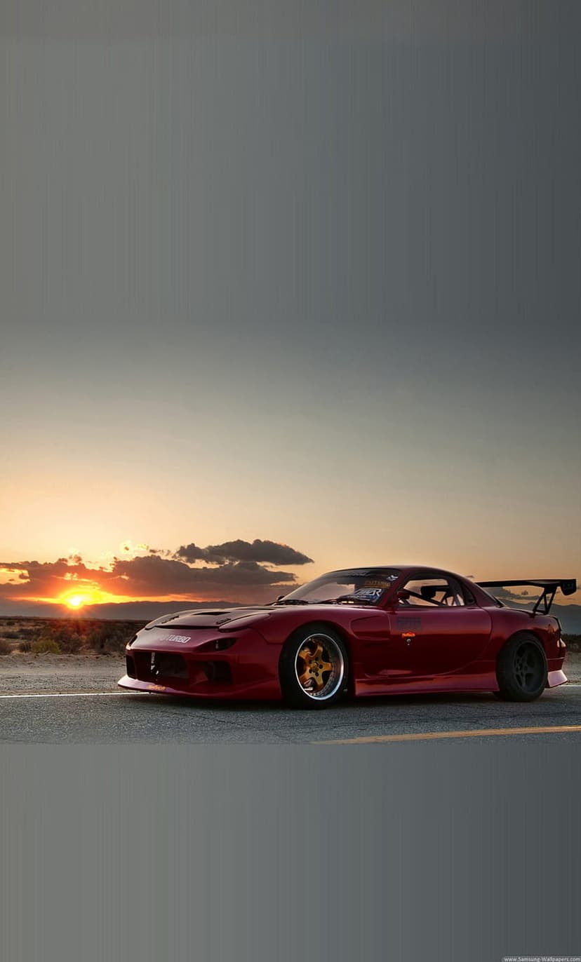 Red Mazda RX-7 sports car with a large spoiler parked on a desert road during a vibrant orange sunset.