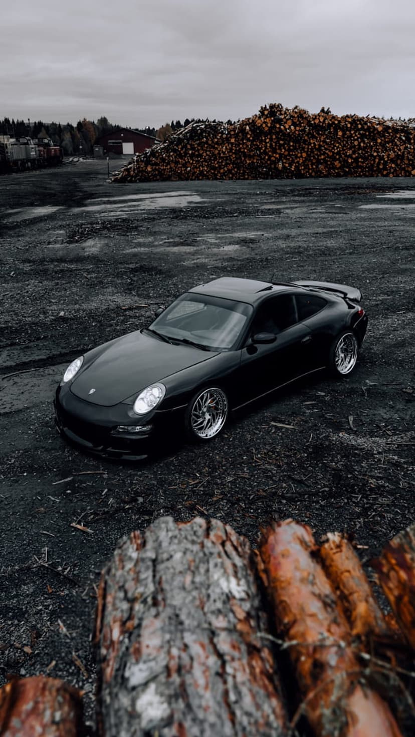 Black Porsche 911 sports car parked on dark gravel in front of a giant stack of logs under a cloudy gray sky.