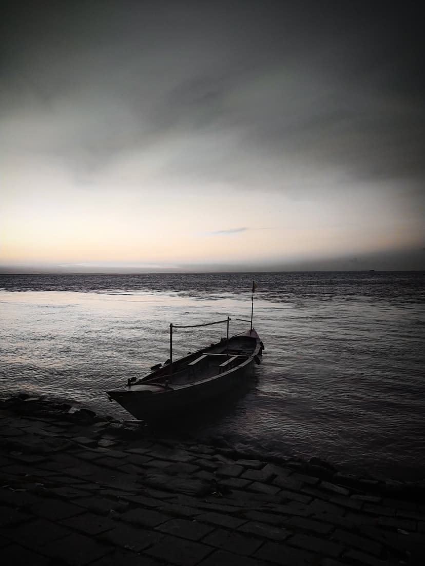 Wooden boat drifts on dark rippling water near a rocky shore under a dim gray twilight sky at the edge of a lake.