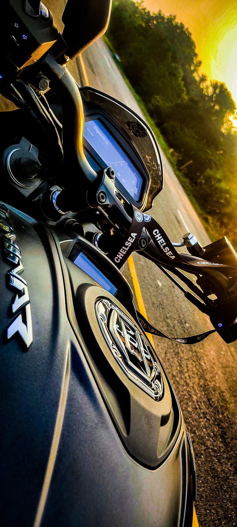Motorcycle handlebars and digital dashboard overlook a sunlit road at dusk with trees in the background.