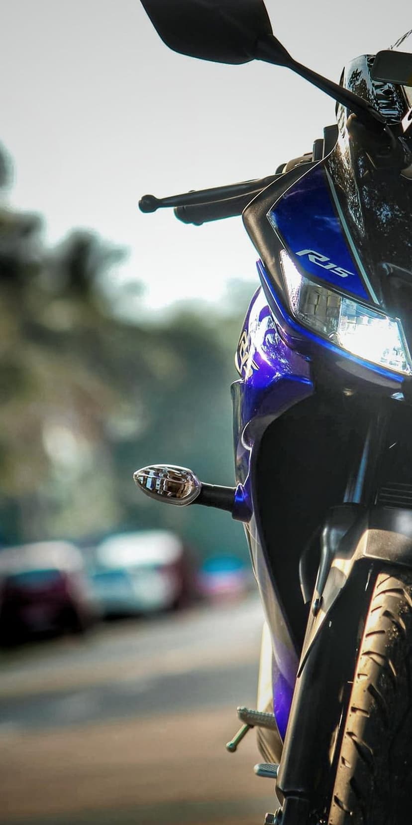 Blue Yamaha R15 motorcycle headlight and textured front tire on a sunny road with blurred cars background.