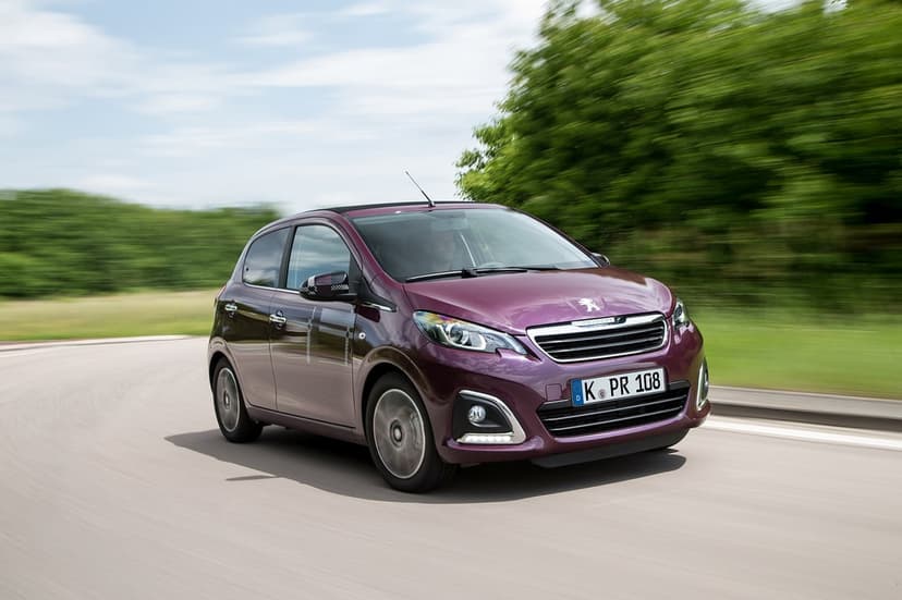 Purple Peugeot 108 hatchback driving fast on a curved road with motion-blurred green trees and a blue sky.