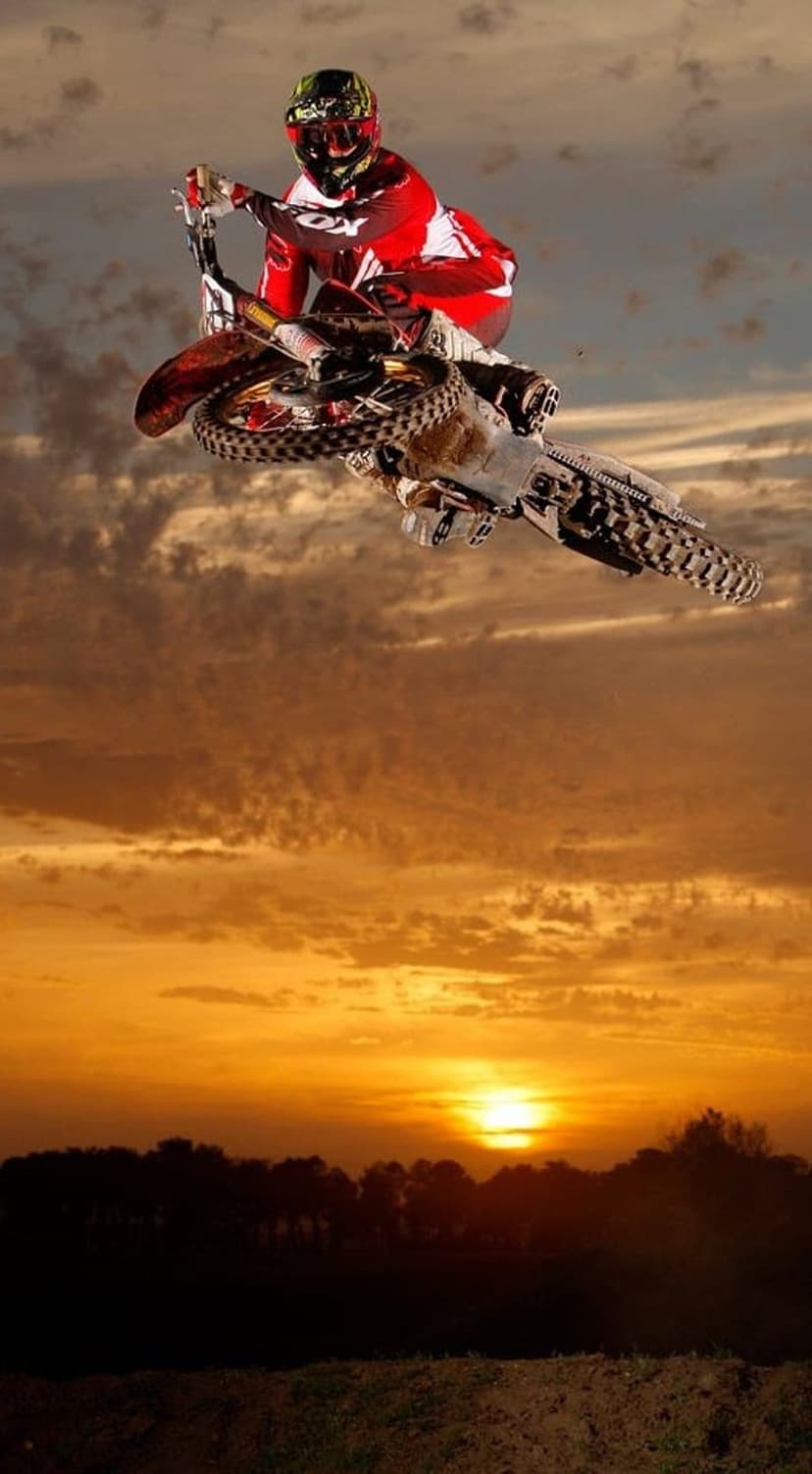 Motocross rider in red gear performs an acrobatic mid-air jump against a vibrant orange and yellow sunset sky.
