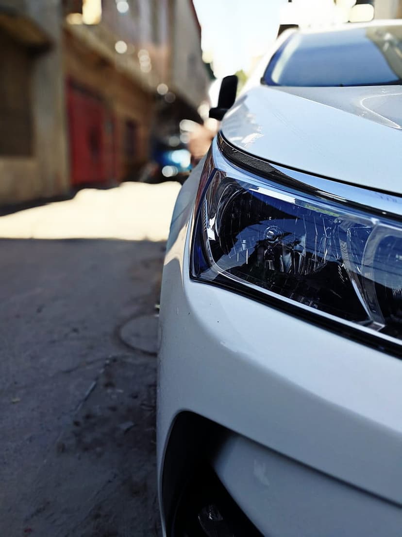 White car headlight with chrome accents and sunlight reflections in a blurred urban alleyway seen from a low angle.