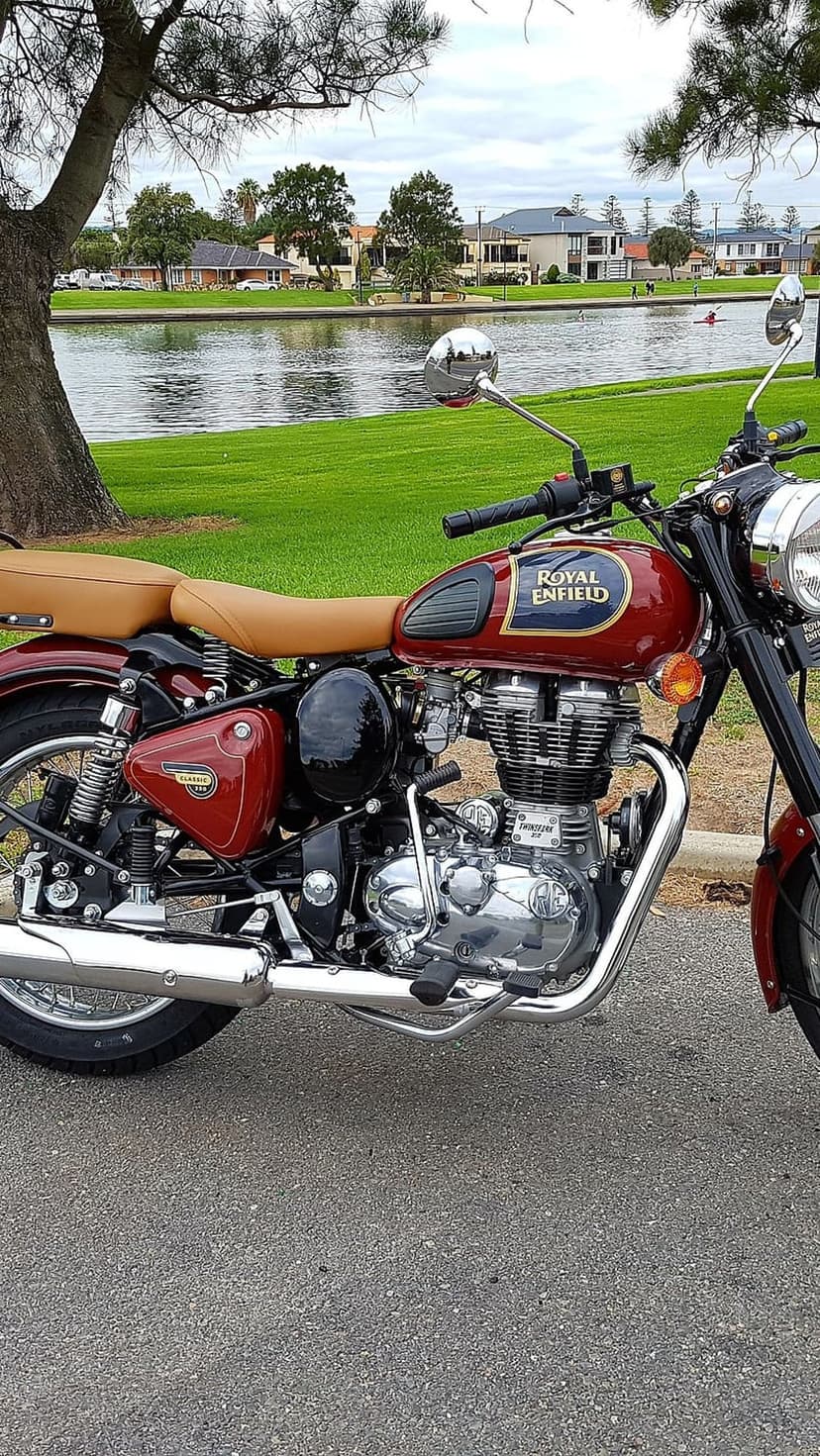 Maroon Royal Enfield Classic bike with a tan seat parked on asphalt near a calm lake under overcast skies.