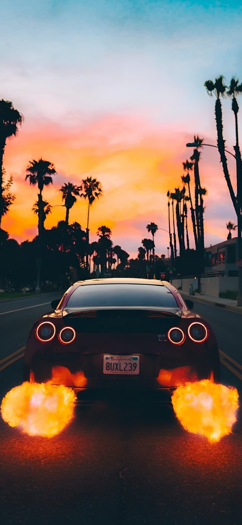 Nissan GT-R emits bright orange exhaust flames on a palm tree lined road during a vibrant tropical sunset.