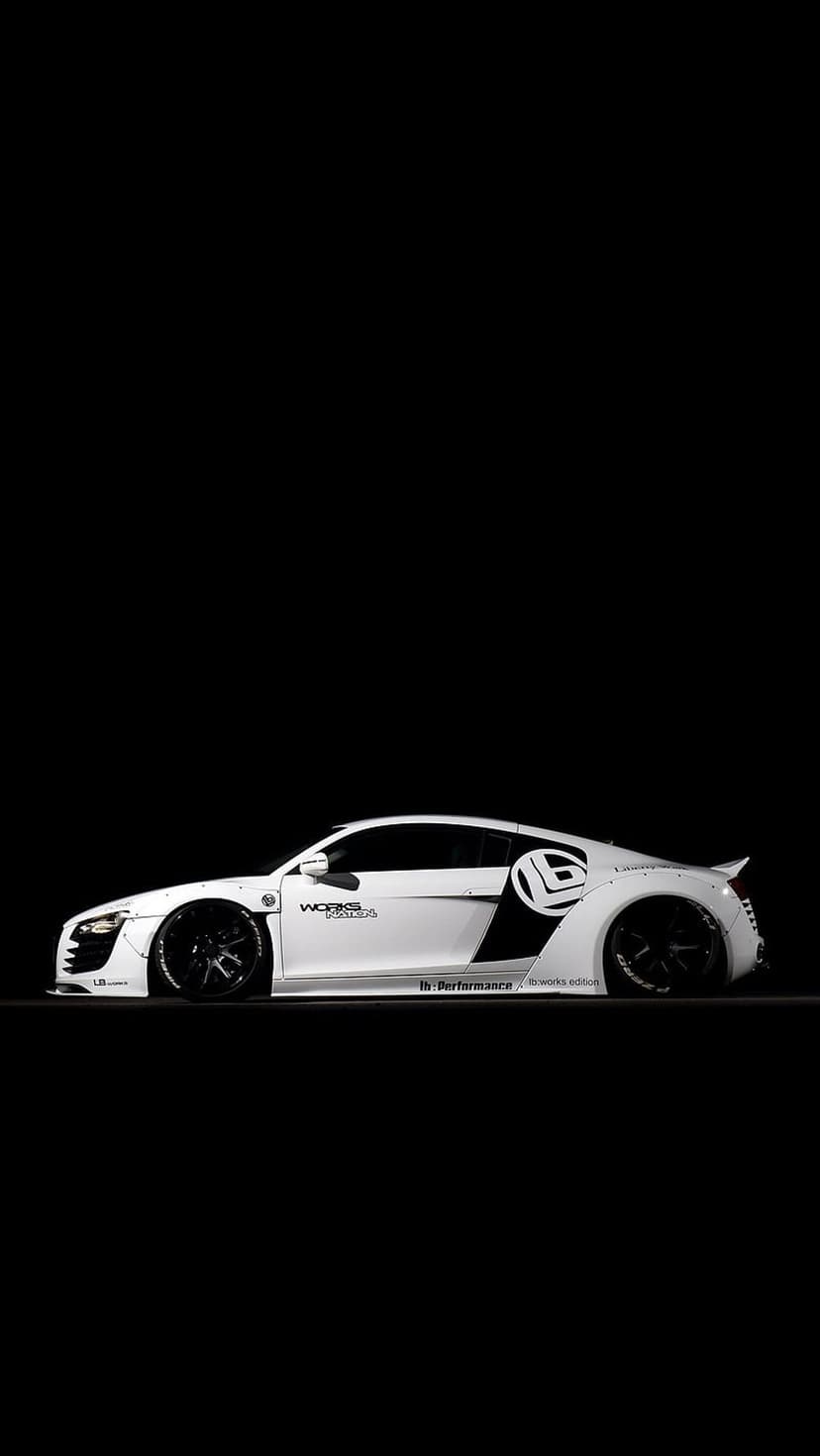 White Audi R8 with widebody fenders and black accents parked in a dark studio under moody spotlighting.