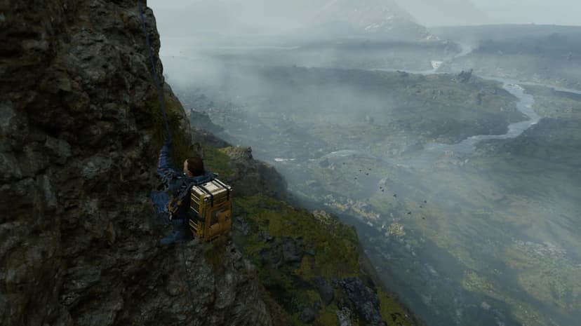 Sam Porter Bridges climbs down a rocky cliff face with a blue rope overlooking a vast, misty river valley.