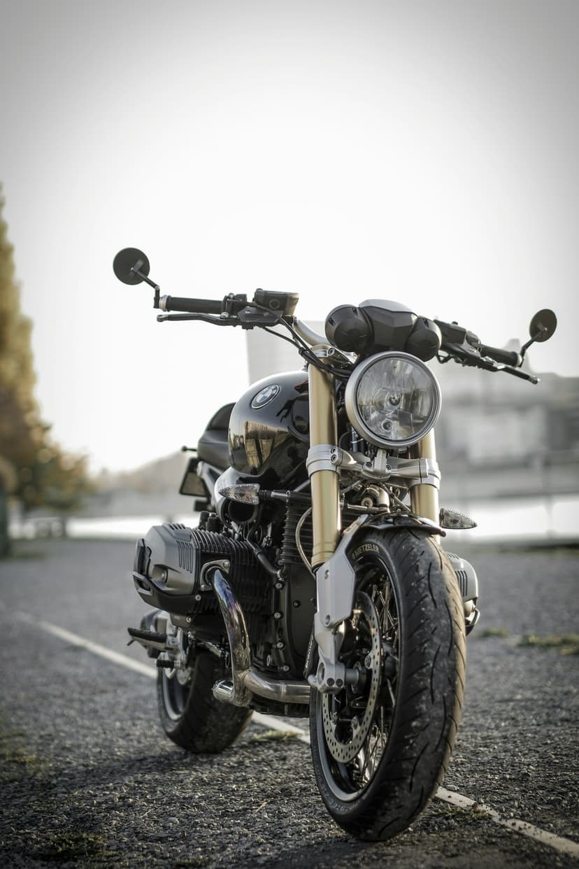 BMW R NineT motorcycle with gold forks and a round headlight parked on a gravel road near a blurred treeline.