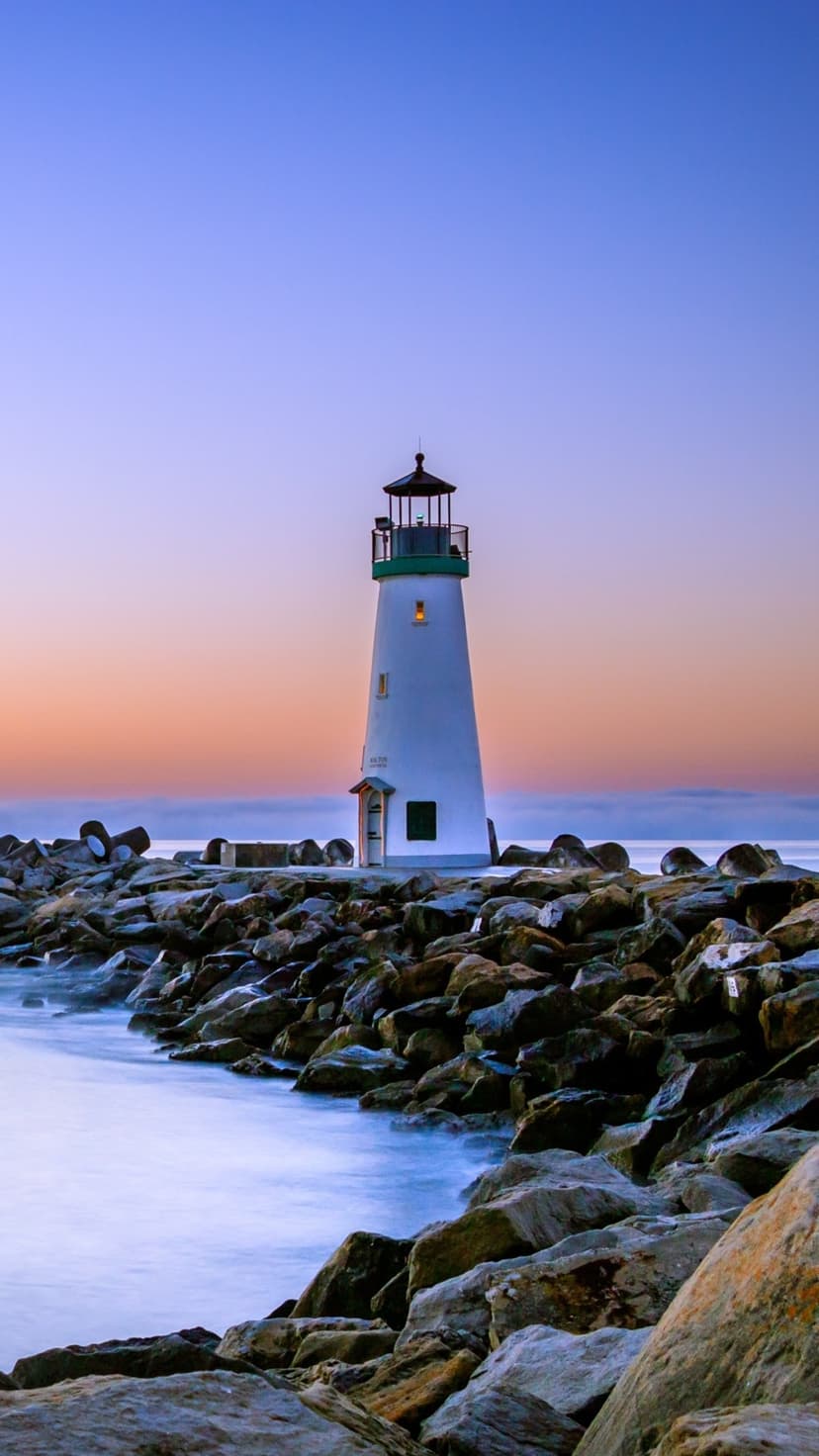 White lighthouse with a green cap stands on a rocky breakwater during a soft orange and blue sunset.