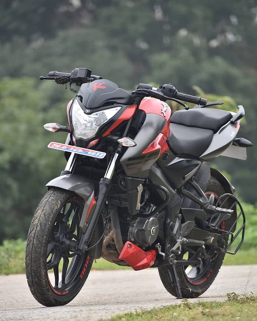 Red and black Bajaj Pulsar NS200 motorcycle parked on asphalt with a blurred green woodland backdrop.