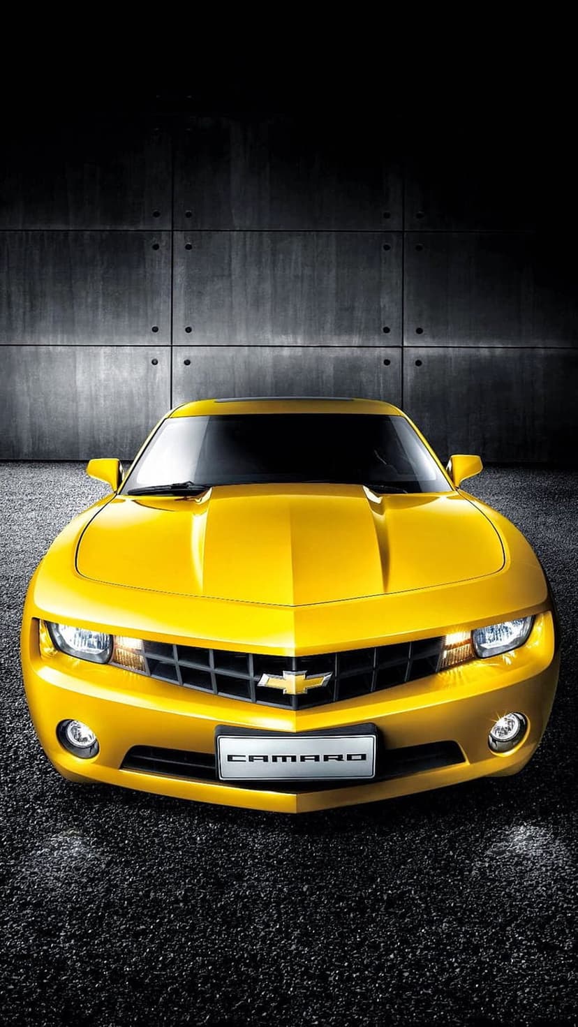 Yellow Chevrolet Camaro parked in front of a dark concrete wall with glowing headlights on a gravel surface.
