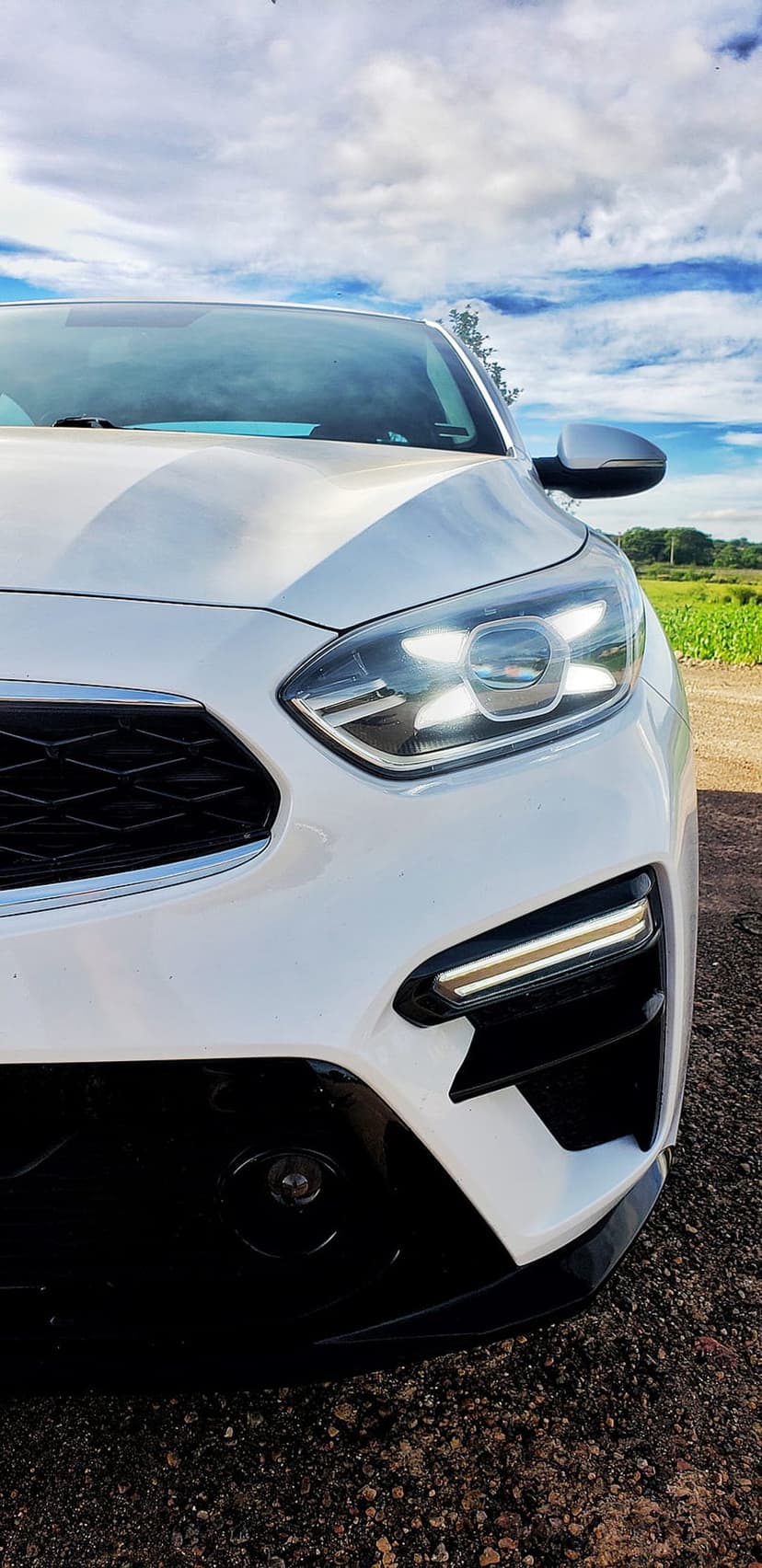 White Kia Forte front grille and LED headlights parked outdoors by a green field under a bright cloudy sky.