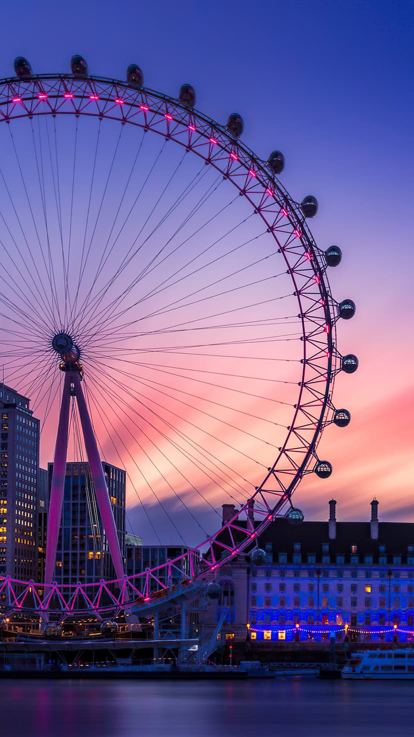 London Eye Ferris wheel silhouetted against a purple and pink sunset sky reflected on the River Thames surface.
