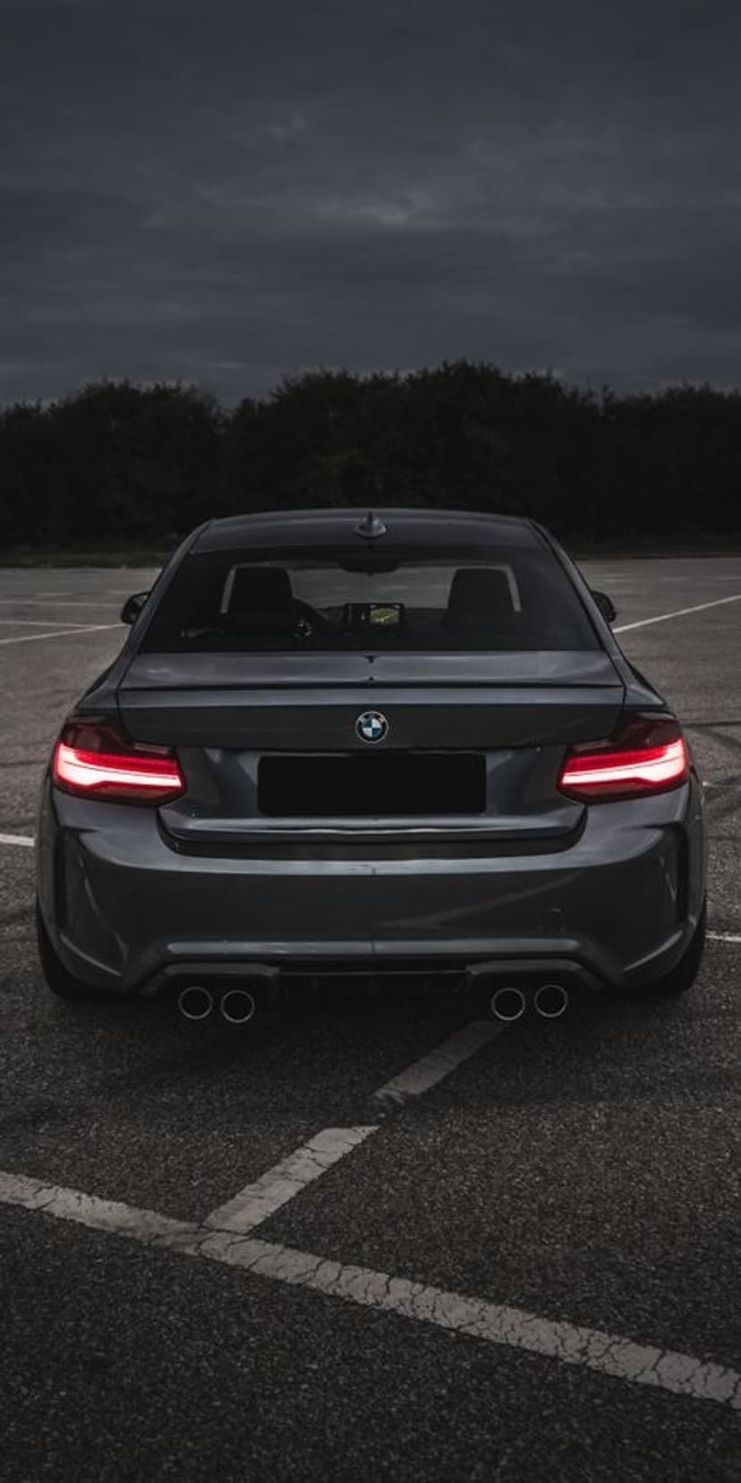 BMW M2 Coupe rear view showing glowing red LED taillights and quad exhausts on dark asphalt under a moody sky.