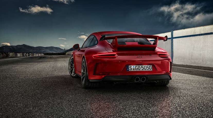 Red Porsche 911 GT3 with a large rear wing parked on an asphalt racetrack under a dramatic cloudy sky.