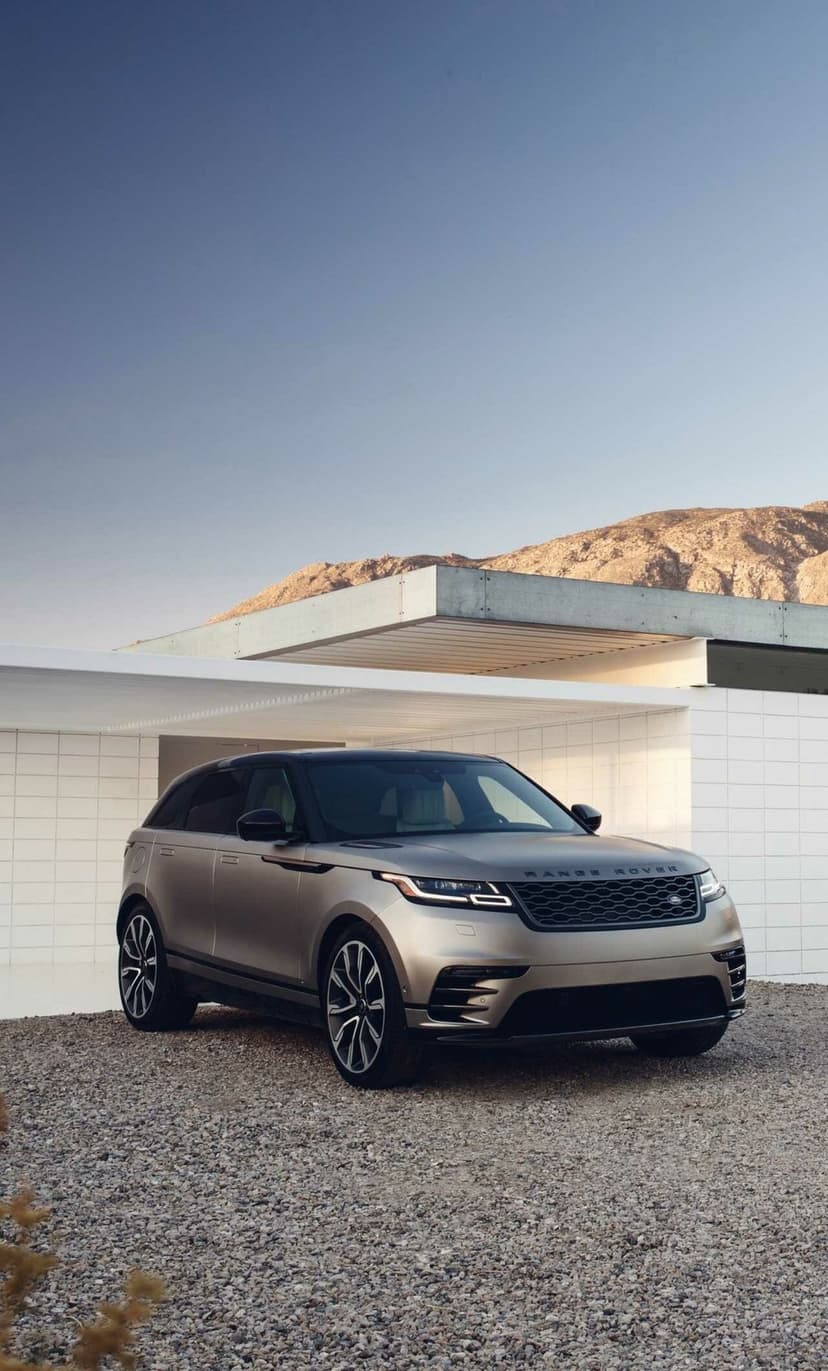 Silver Range Rover Velar parked on gravel before a white tiled modern building under a clear blue sky.