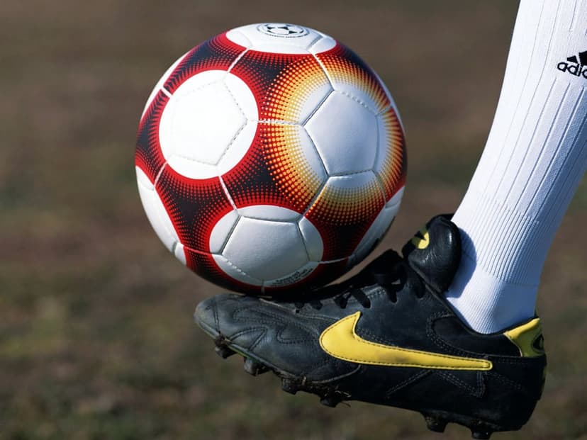 Soccer ball balanced on the tip of a yellow and black Nike cleat with a white Adidas sock on a blurred field.