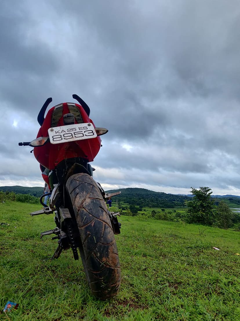Red motorcycle parked on a lush green ridge overlooking forested hills and a cloudy horizon from a rear angle.