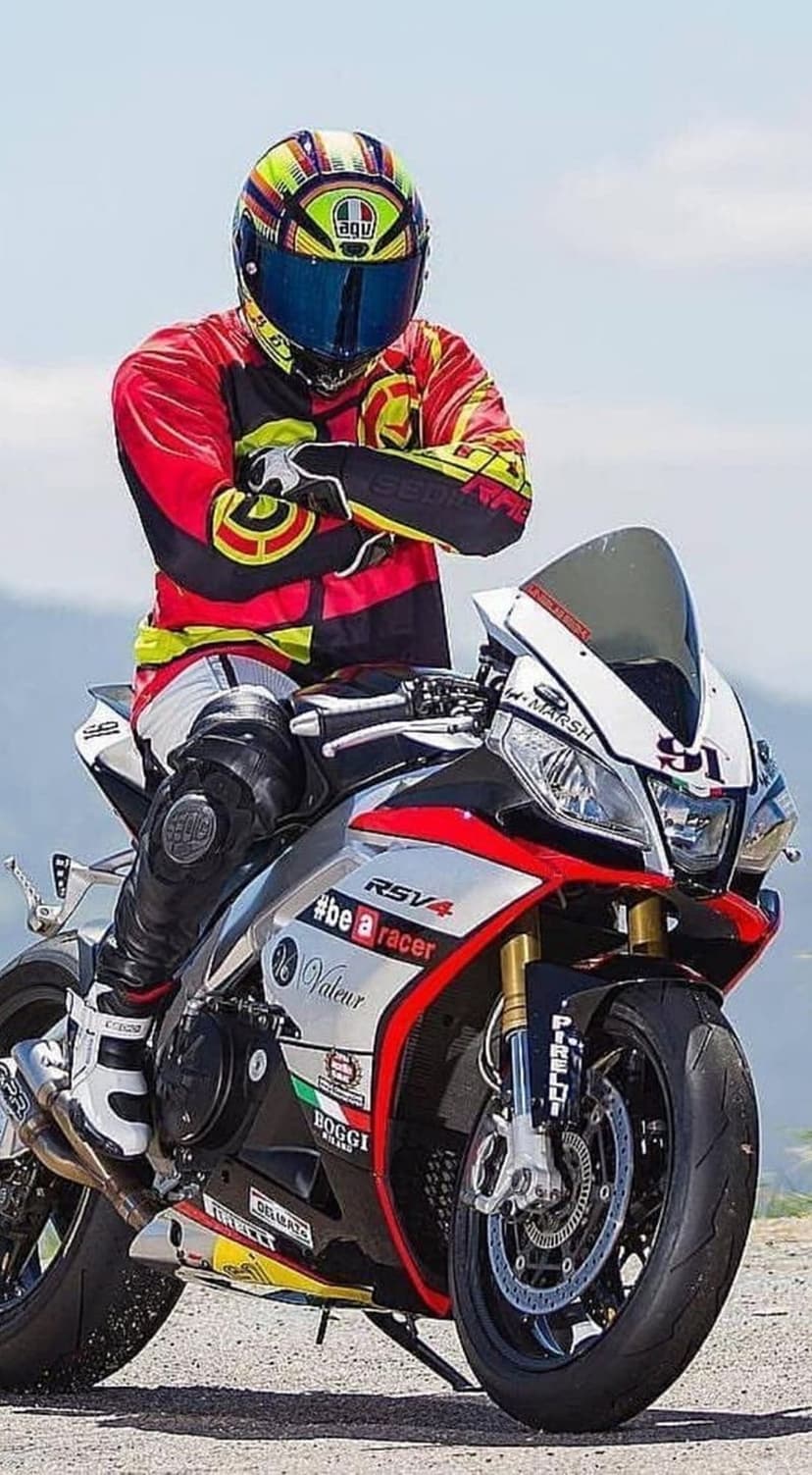 Motorcyclist in a red racing suit and AGV helmet leans against a white and red Aprilia RSV4 sportbike.