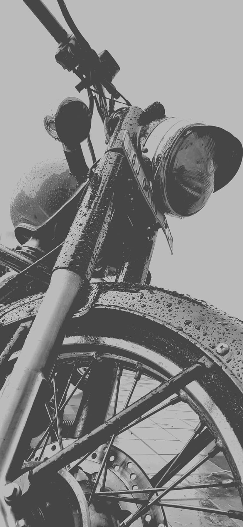 Vintage motorcycle front end covered in rain droplets with visible spokes and headlight in a grayscale palette.