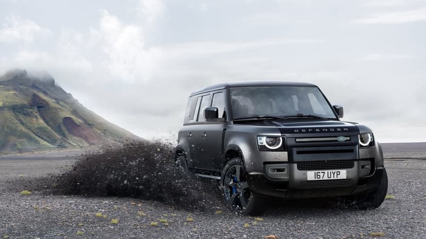 Black Land Rover Defender kicks up gravel on a volcanic plain with a misty mountain under a cloudy sky.